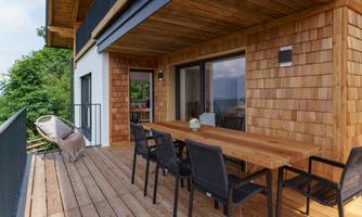 A modern balcony with a wooden terrace and a large dining table. The surroundings are green and inviting.