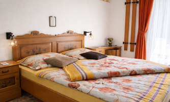 A cozy bedroom with a large bed and colorful floral bedding. The warm wooden furniture and the orange curtain create an inviting atmosphere.