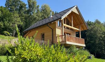 A beautiful wooden house with a balcony and a clear blue sky. Surrounded by greenery and trees.