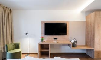 A modern bedroom with a wooden desk and a large television. The decor includes a cozy armchair and a small bouquet of flowers.