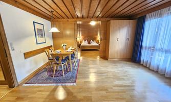 A bright room with wooden floor and wooden ceiling. There is a dining area with table and chairs as well as an adjacent bedroom.