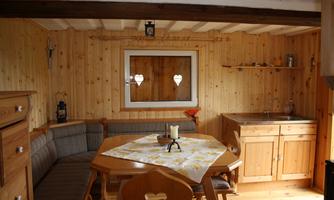 A cozy wooden interior with a dining area and a small kitchen. The table is decorated with a tablecloth and there is a window with heart motifs.