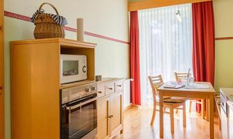 A bright kitchen with wooden shelves and large windows. A dining table with chairs and modern kitchen appliances are visible.