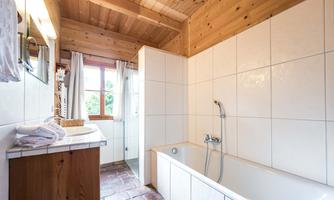 A bathroom with wooden ceilings and white tiles. There is a bathtub, a sink, and windows that let in natural light.