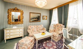 An elegant living room with floral furniture and a large mirror. The walls are decorated with stylish wallpaper, and there is a table with a fruit bowl in the center.