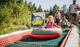 A little girl is happily sitting in a tire and preparing for a slide down in front of a slide. In the background, there are more people and a sign with the logo of a theme park.