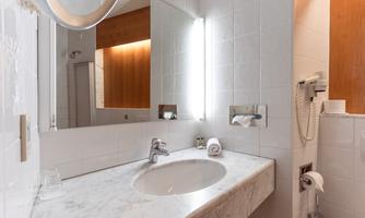 A modern bathroom with white tiles and a large mirror. On the marble countertop are towels and toiletries.