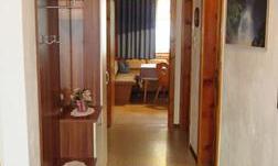 A narrow hallway with a wood ceiling and walls in light colors. On the left, there is a small cupboard and in the background, a table and chairs are visible.