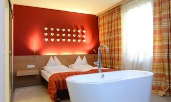 A cozy hotel room with a double bed and a freestanding bathtub. The walls are painted in warm colors, and large windows ensure plenty of light.