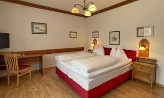 A cozy bedroom with a large, red bed and two pillows. There is a wooden desk and chairs, as well as warm lighting.