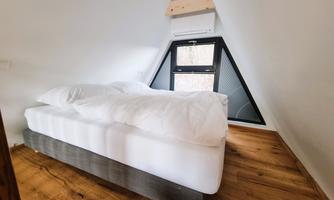 A cozy bedroom with a large bed and bright wooden floor. The window is designed in a modern, triangular shape.