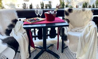 A cozy balcony with two chairs and a table. On the table are glasses, a bowl of drinks, and a fruit platter, surrounded by snow-covered trees.