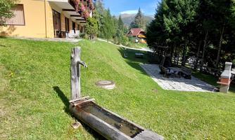 A tranquil garden with a wooden fountain. In the background, there are trees and a terrace with seating.