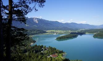 A picturesque landscape with a clear lake, surrounded by mountains and green forests. In the foreground, some buildings are visible that lie by the shore.