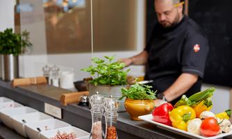 A chef is preparing fresh ingredients in a modern kitchen. Various vegetables and spices are visible on the table.