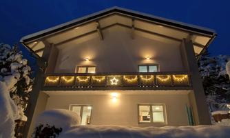 A cozy house in the snow, illuminated by festive lights. The evening sky is dark blue and the surroundings are wintery and snowy.