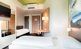 A modern hotel room with two beds, a table, and a television. The walls are brightly designed and there is plenty of natural light.