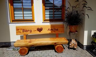 A wooden bench with the inscription "Berg ♥ Arche" stands in front of a building. Next to the bench is a planter with decorative plants.