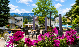 A beautiful garden with colorful balcony flowers and tables. In the background, there are mountains and a swimming pool.