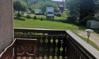 A balcony with a wooden railing and cushions. In the background, meadows and trees can be seen.