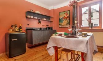 A modern kitchen with pink walls and wooden flooring. The table is set with dishes and wine glasses, and there are open shelves with kitchen utensils.