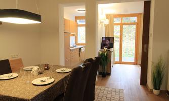 A modern living and dining room with a large table and chairs. In the background, there is a kitchen and access to the garden.