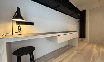 A modernly furnished workspace with a simple table and a black stool. A stylish lamp provides lighting in a bright room.