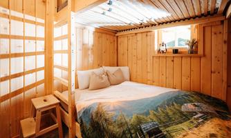 A cozy wooden bedroom with a loft bed and a colorful bedspread. Light streams through the window onto the simple furnishings.