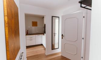 A bright hallway with a large mirror and a white door. Modern kitchen furniture and a warm wood floor can also be seen.