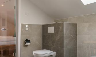 A modern bathroom with gray tiles and a toilet area. The room has a large window that lets in daylight.