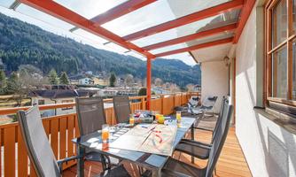 A sunny balcony with a large table and comfortable chairs. In the background, green mountains and a clear sky can be seen.