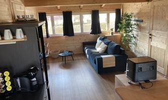 A cozy room with wooden walls and large windows. In the foreground, there is a sofa, a table, and a small kitchenette.