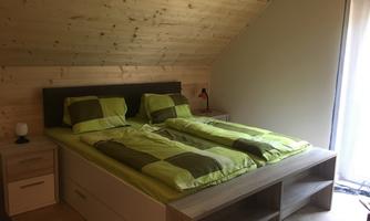 A cozy bedroom with a double bed and green bedcovers. The walls are made of wood, and there is a nightstand as well as a window with curtains.