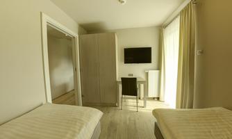 A simple room with two single beds, a desk, and a wall-mounted television. Large windows provide plenty of natural light.