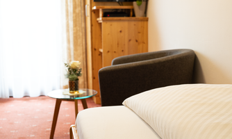A cozy room with a bed, an armchair, and a small table. The room has large windows and a TV.