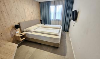 A modern hotel room with two single beds and light wooden decor. The room has a window with curtains and a television on the wall.