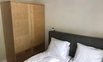 A cozy bedroom with a double bed and two pillows. A wooden wardrobe stands against the wall and a small clock is visible.