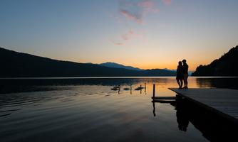 A romantic couple stands by the shore of a tranquil lake at sunset. Gentle colors reflect in the water, creating a peaceful atmosphere.