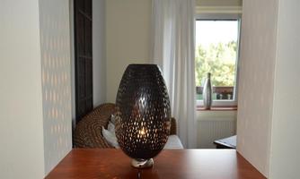 A decorative lamp stands on a wooden shelf. In the background, you can see a cozy living area with a window and curtains.