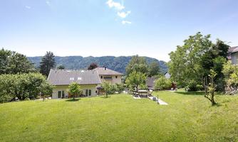 A quiet garden with green grass and several trees. In the background, mountains and a clear sky can be seen.