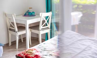 Bright dining area with a white table and two chairs, decorated with turquoise glasses – right next to the balcony door, allowing plenty of light and nature to flow into the room.