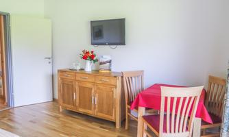 A cozy living space with wooden floors and a table with red tablecloths. A TV hangs on the wall, and there is a sideboard decorated with flowers.