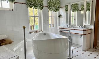 A modern bathroom with an elegant bathtub and large windows. The walls are light tiled, and the curtains have a beautiful green pattern.
