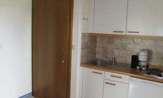 A small kitchen with a wooden door and white cabinets. The equipment includes a sink, a coffee machine, and a refrigerator.