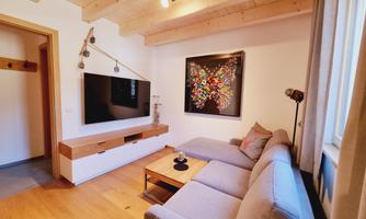 A modern living room with a gray couch and a wooden table. A colorful artwork hangs on the wall and a television stands opposite.