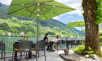 A picturesque café by the lake with green sun umbrellas and a view of the mountains. Two guests enjoy the scenery outdoors.