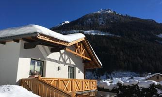 A cozy wooden house in the snow with an impressive mountain in the background. The sky is clear and blue, perfect for winter activities.