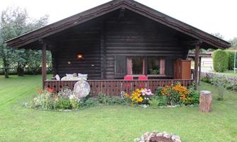 A cozy wooden house with a green garden. Surrounded by colorful flowers and a small fire pit in the foreground.
