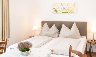 A cozy bedroom with a double bed and two large pillows. On the table, there is a plant, and a colorful picture hangs on the wall.