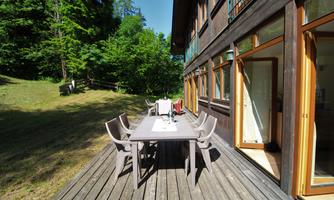 A cozy terrace area with a large table and several chairs. In the background, green trees and a sunny sky are visible.